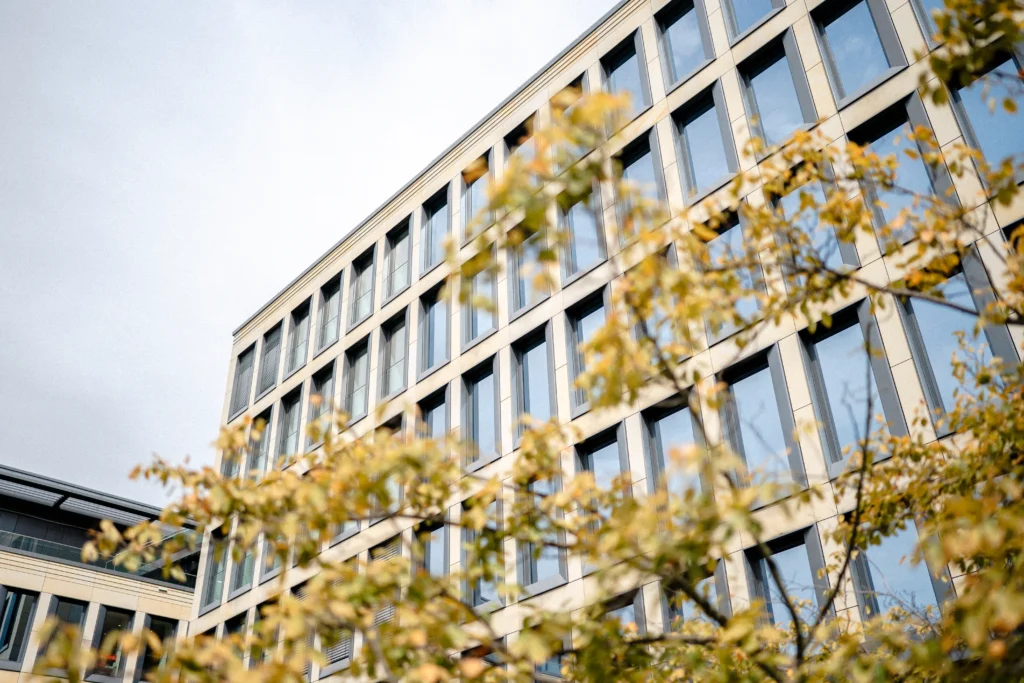 Moderner Bürokomplex mit großen Fensterflächen, fotografiert durch die Äste eines Baumes im Vordergrund.