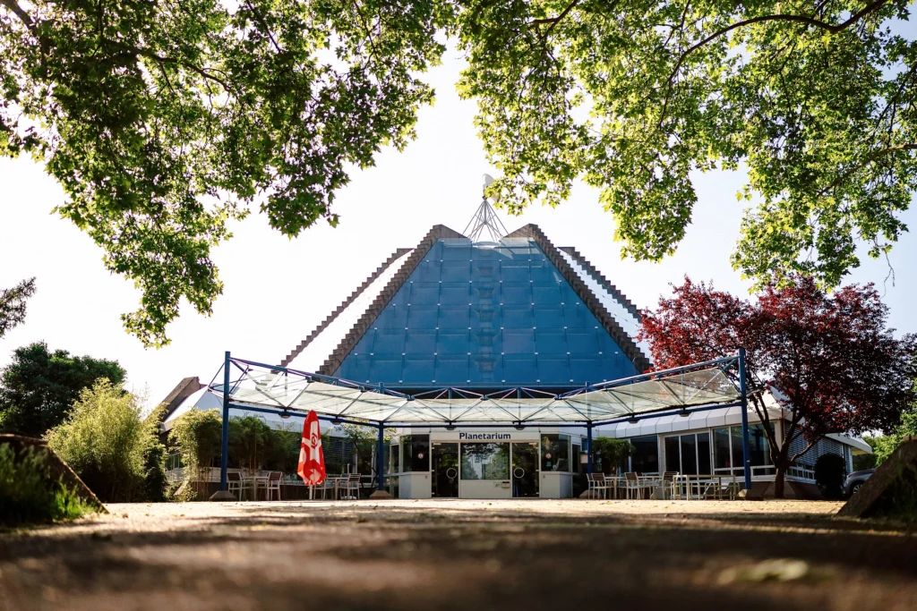 Das Planetarium mit seiner gläsernen Pyramidenform steht hinter einem überdachten Eingangsbereich und wird von großen Bäumen eingerahmt.