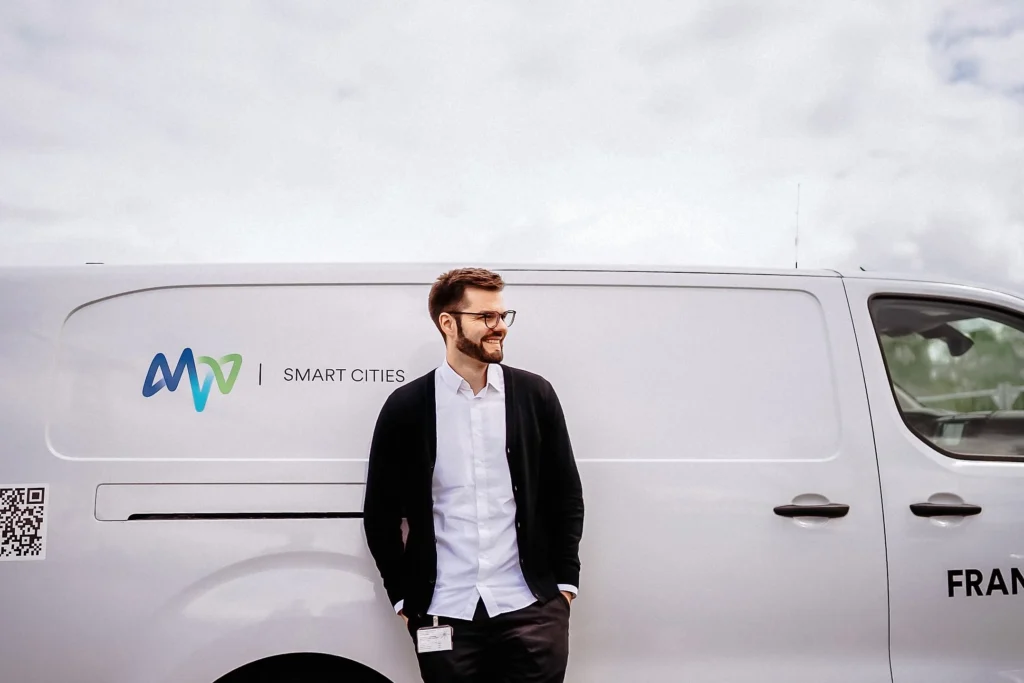 Eine Person lehnt lächelnd vor einem Transporter mit dem Schriftzug „MVV Smart Cities“.