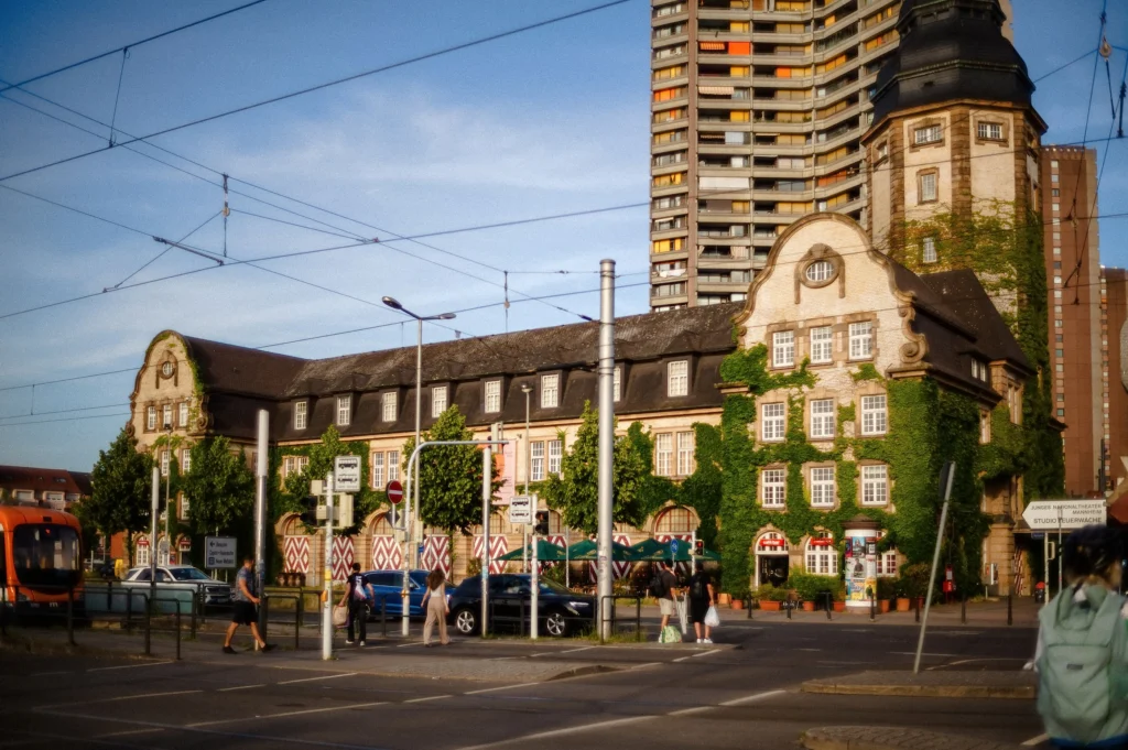 Historisches Gebäude der Alten Feuerwache Mannheim mit verziertem Fassadenteil und hohem Wohnhochhaus im Hintergrund an einer Straßenkreuzung mit Straßenbahn und Fußgängern.