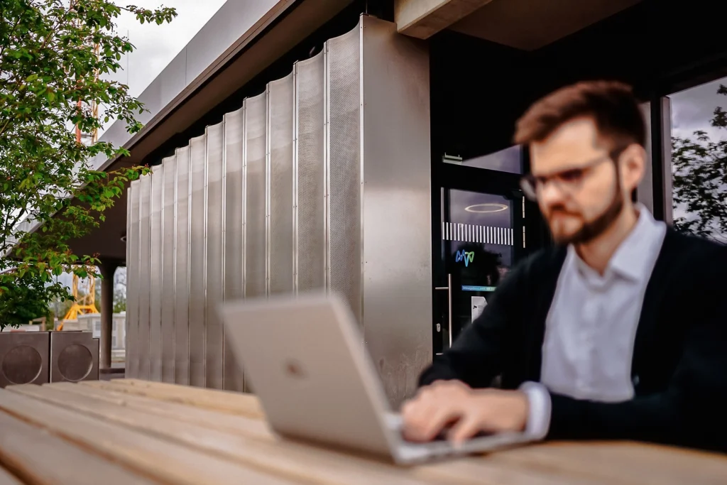 Eine Person arbeitet mit einem Laptop an einem Tisch vor einem modernen Gebäude.