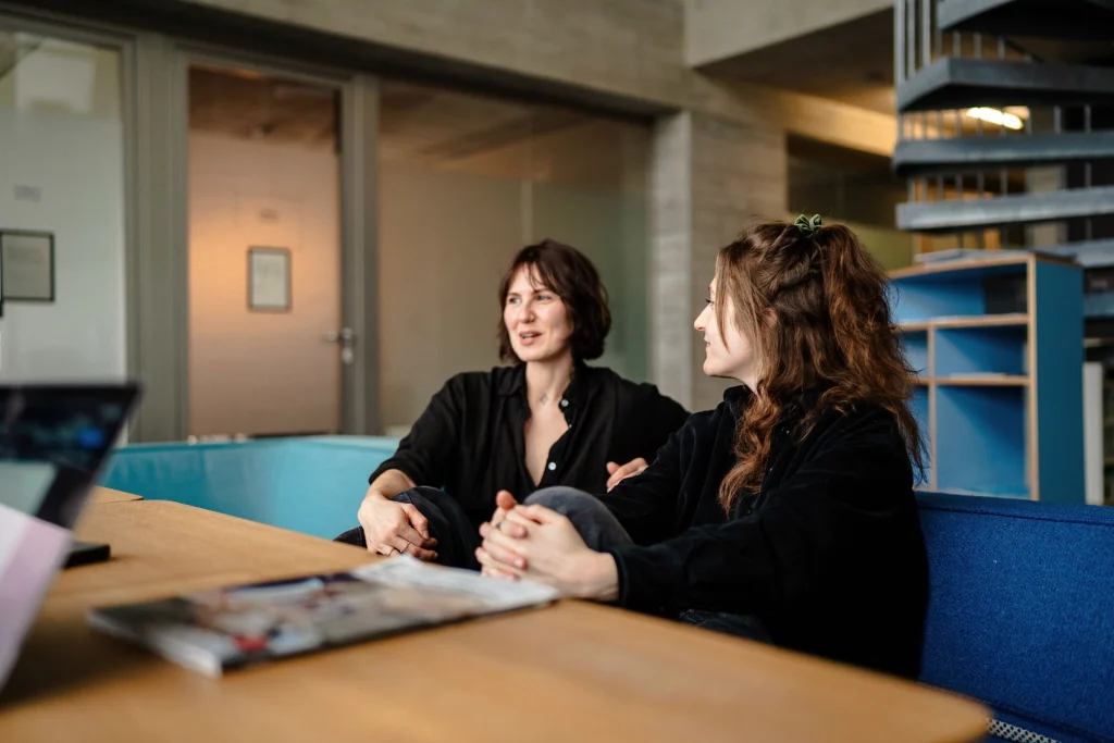 Zwei Personen sitzen in einem modernen Büro auf einer Sitzbank und unterhalten sich an einem Tisch.