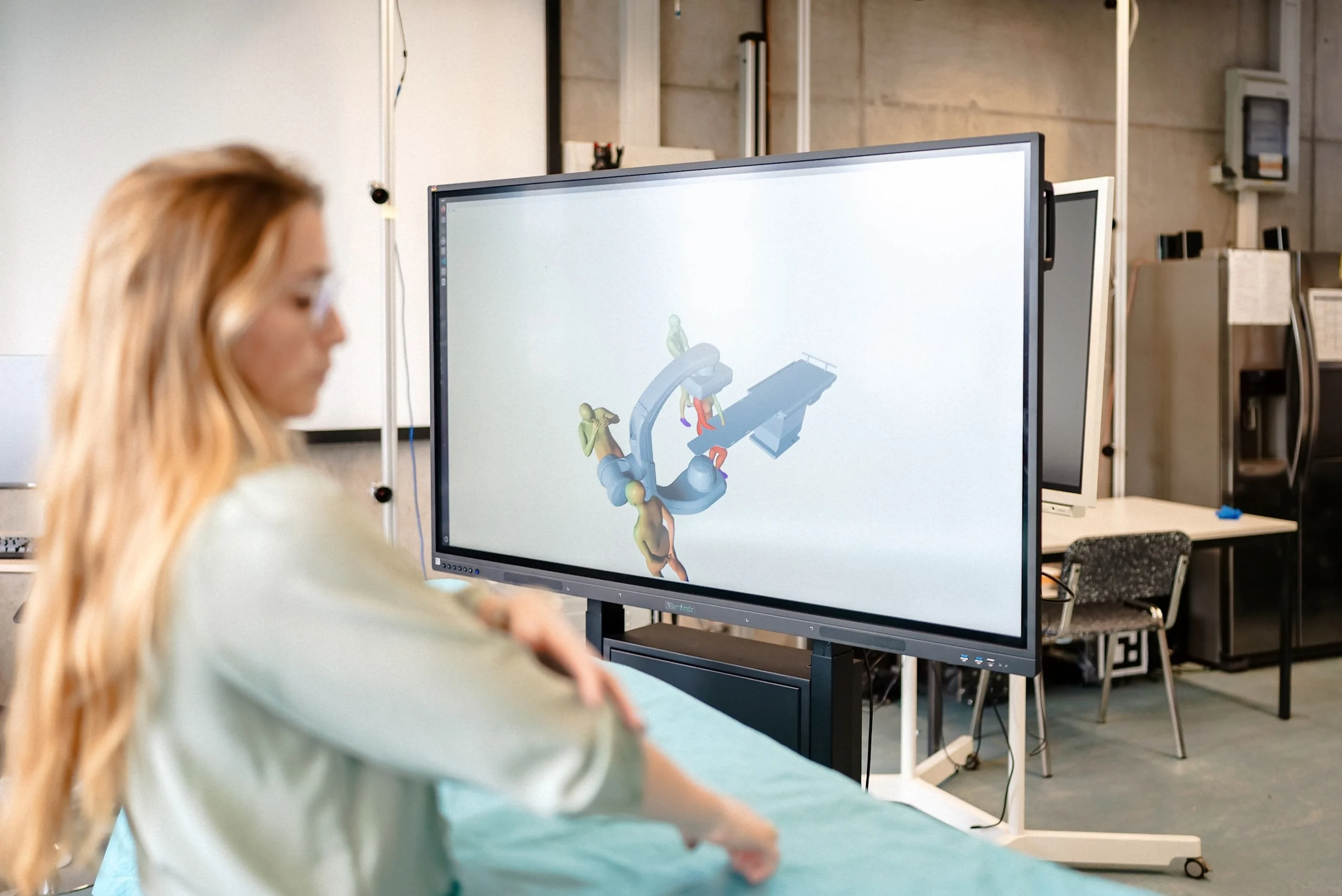Eine Person betrachtet in einem technischen Labor eine 3D-Simulation eines medizinischen Geräts auf einem großen Bildschirm neben einem abgedeckten Untersuchungstisch.