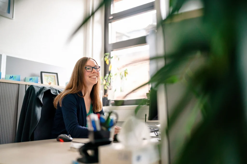 Eine Frau sitzt lächelnd an ihrem Schreibtisch in einem hellen Büro und arbeitet am Computer.