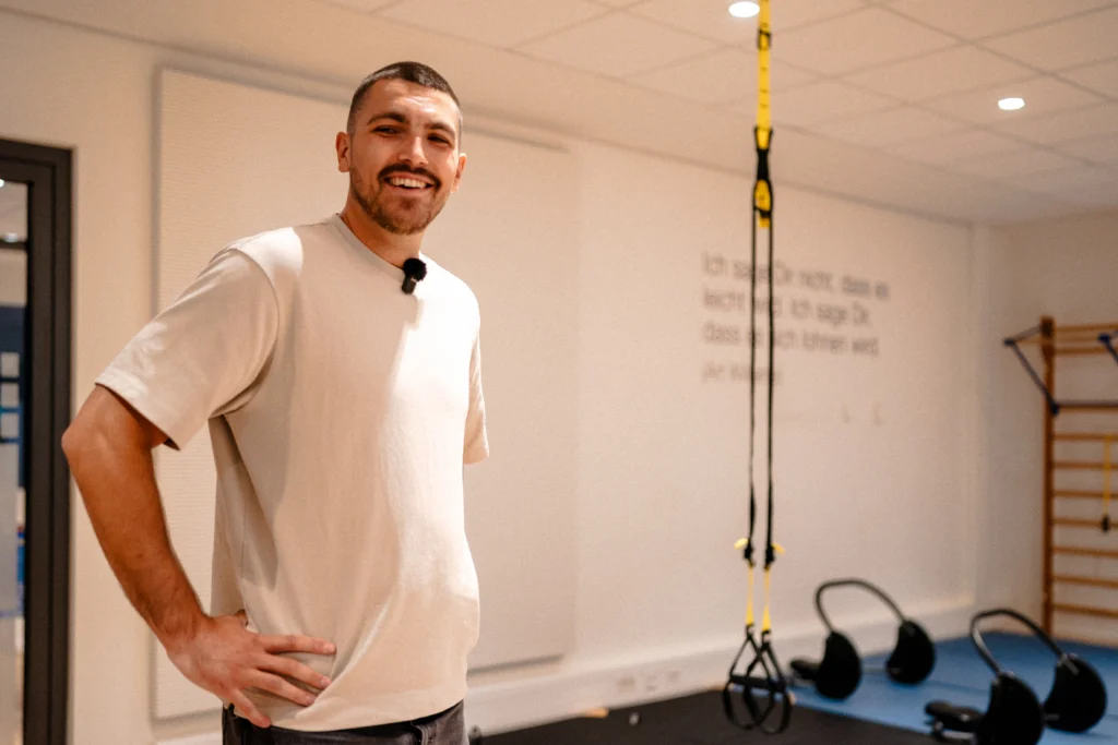 Person steht in einem Fitness- und Bewegungsraum mit Trainingsgeräten und Schlingentrainern im Hintergrund.