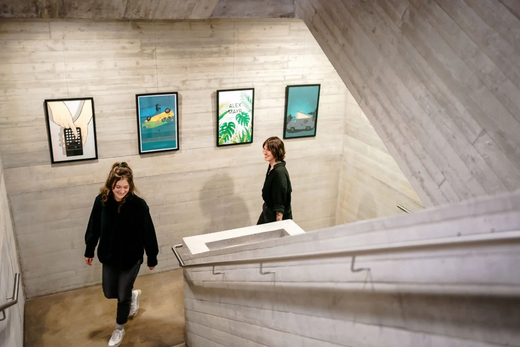 Zwei Personen gehen eine Treppe in einem modernen Bürogebäude hinauf, an einer Wand mit gerahmten Illustrationen vorbei.
