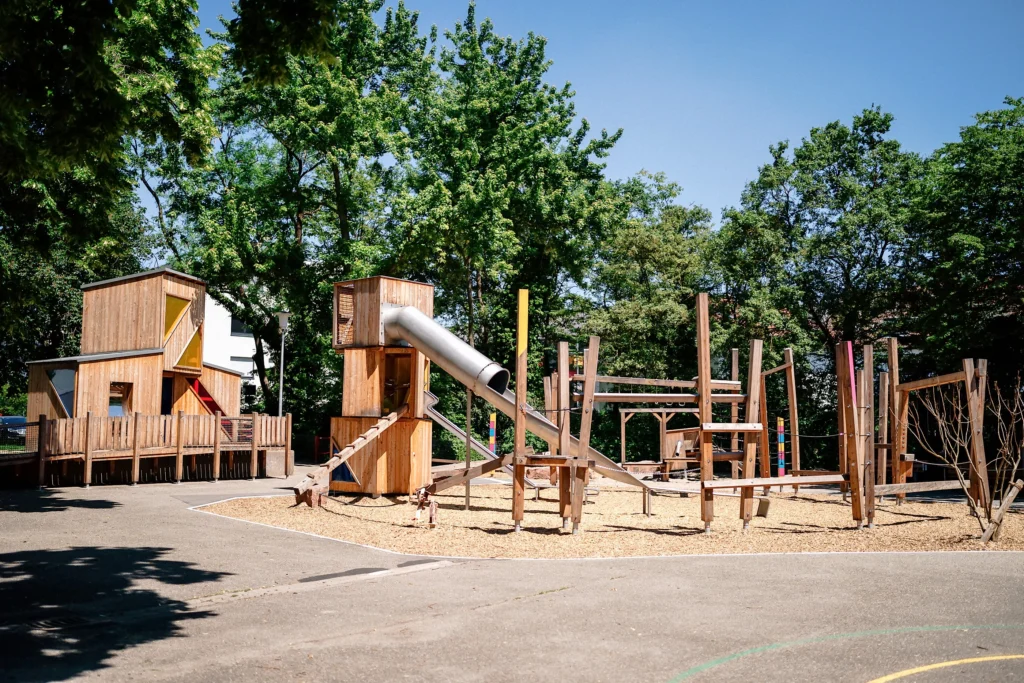 Moderner Holzspielplatz mit Rutsche, Kletterelementen und großzügigen Sitzflächen unter schattenspendenden Bäumen an einem sonnigen Tag.
