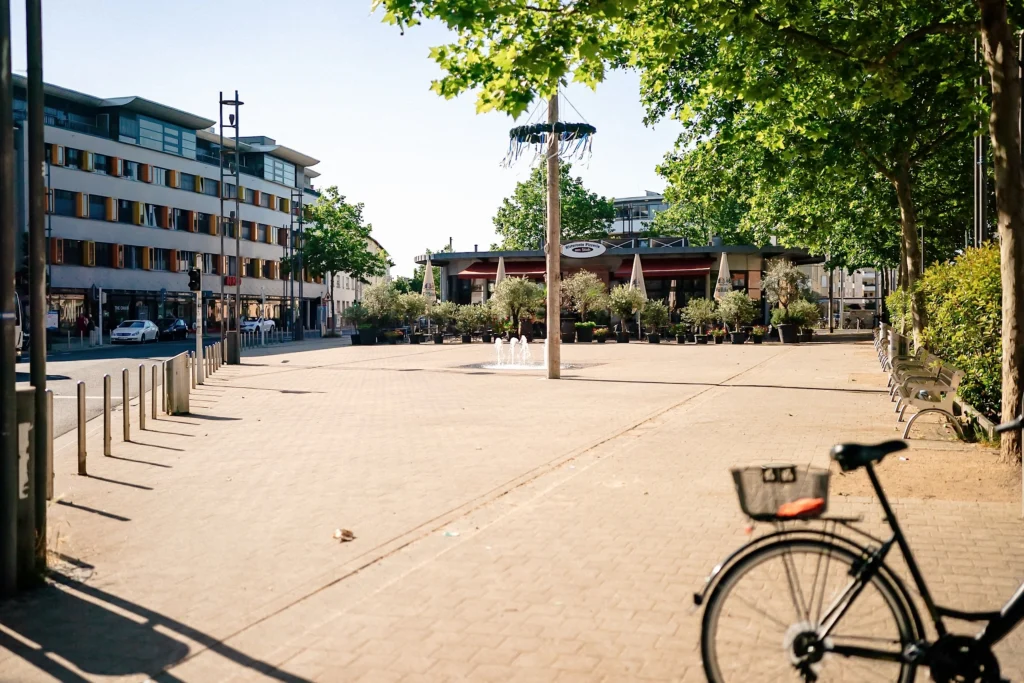 Platz mit Fahrrad, Brunnen und Außengastronomie an einem sonnigen Tag.