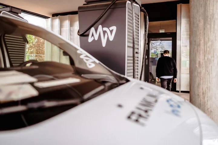 Front part of a white vehicle with lettering in front of a building entrance where a man in a black jacket stands.