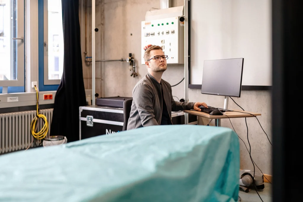 Eine Person sitzt an einem Computerarbeitsplatz in einem technischen Labor neben einem abgedeckten Untersuchungstisch.