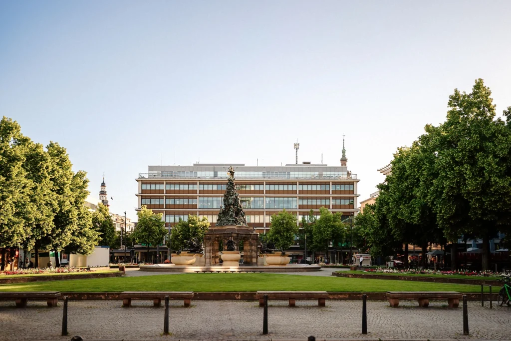 Blick auf den Mannheimer Paradeplatz mit Brunnenanlage und grünen Rasenflächen vor einem mehrstöckigen Kaufhaus an einem sonnigen Tag.