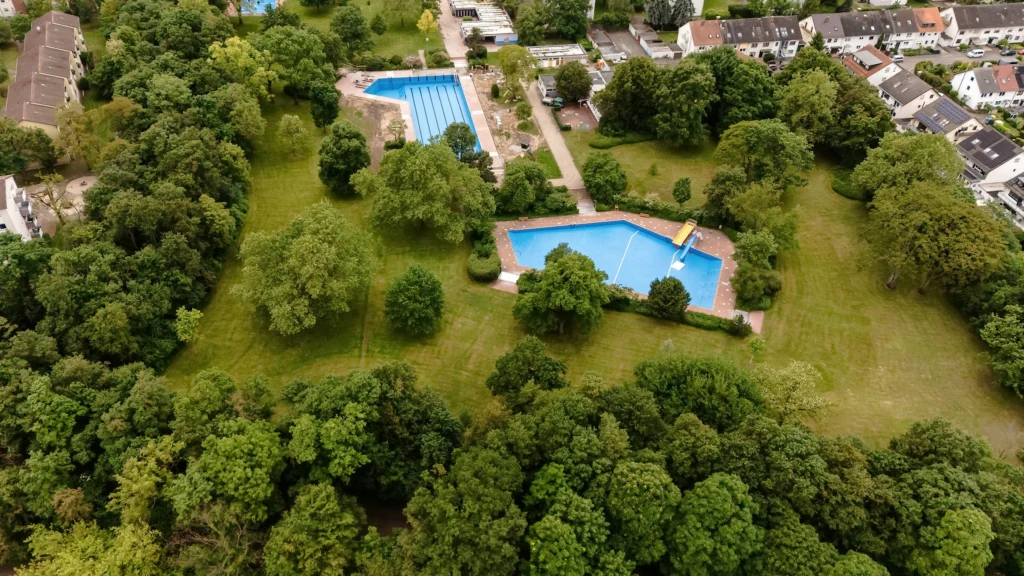 Luftaufnahme des Parkschwimmbads Rheinau mit zwei Schwimmbecken, umgeben von Bäumen und Wohnhäusern.