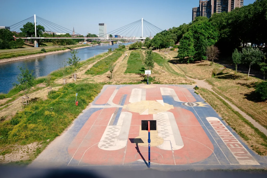 Basketballplatz am Neckarufer in Mannheim mit Blick auf den Fluss, Grünflächen und eine Brücke im Hintergrund.