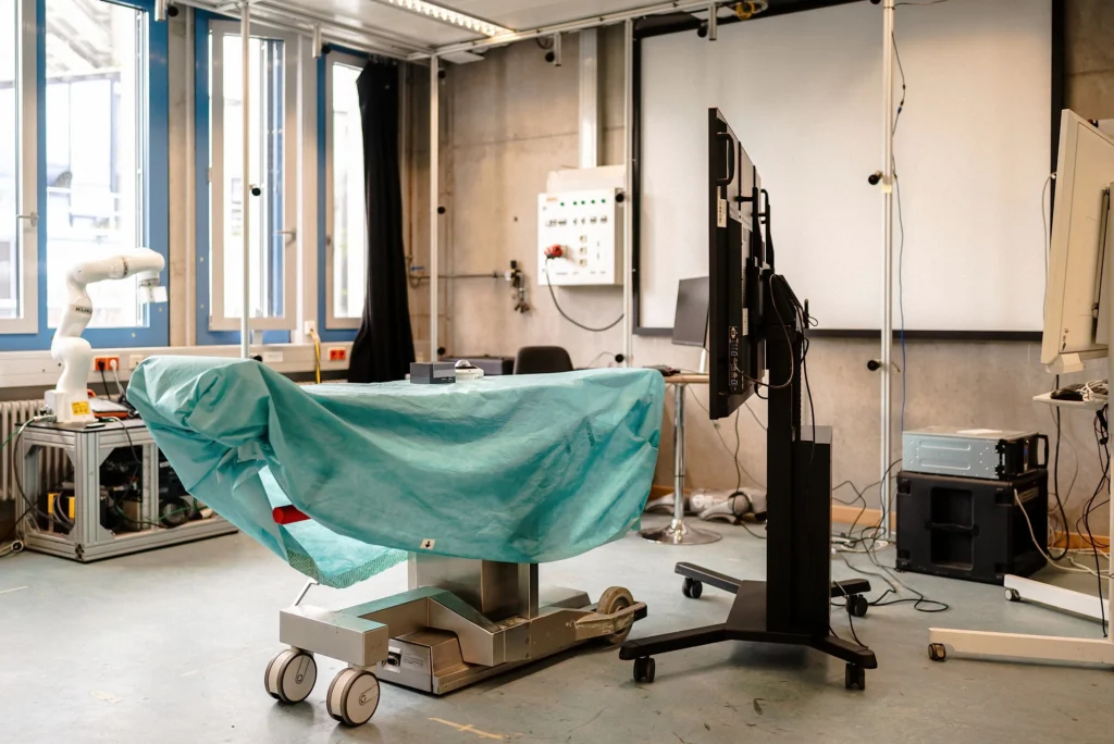 Ein technisches Labor mit einem abgedeckten Untersuchungstisch, einem Roboterarm und mehreren Bildschirmen.