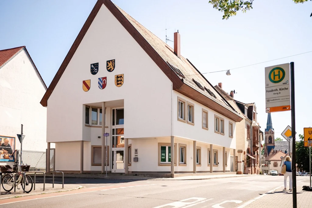 Gebäude im Ortskern von Feudenheim mit Bushaltestelle und Kirchturm im Hintergrund an einem sonnigen Tag.