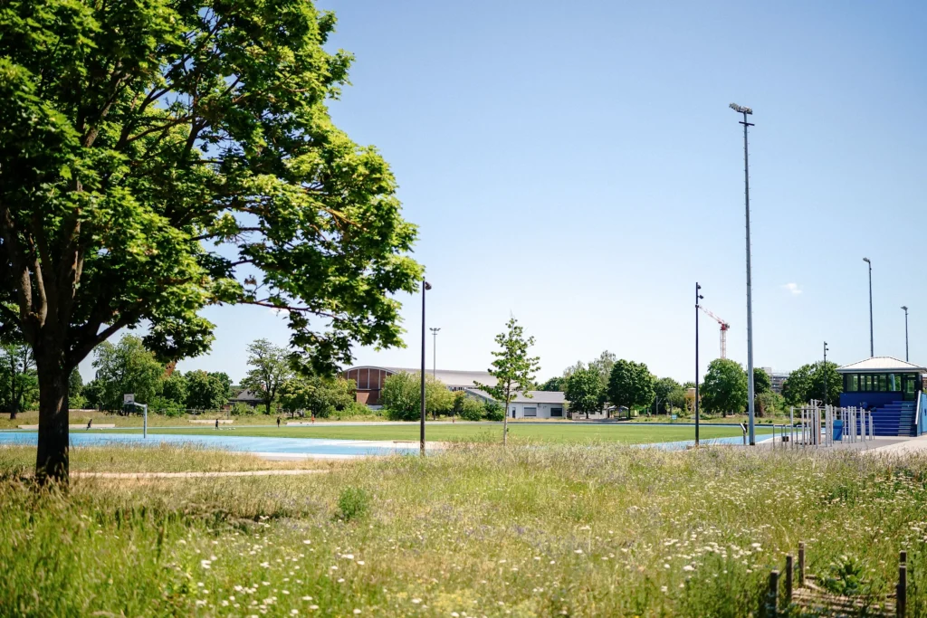 Eine Sportanlage mit Laufbahn und Spielflächen ist von Wiesen, Bäumen und mehreren Gebäuden umgeben.