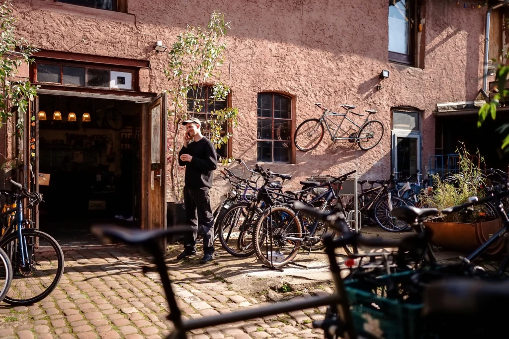 Vor einer Fahrradwerkstatt mit offenstehender Tür stehen zahlreiche Fahrräder im Innenhof, während eine Person im Sonnenlicht vor dem Gebäude zu sehen ist.