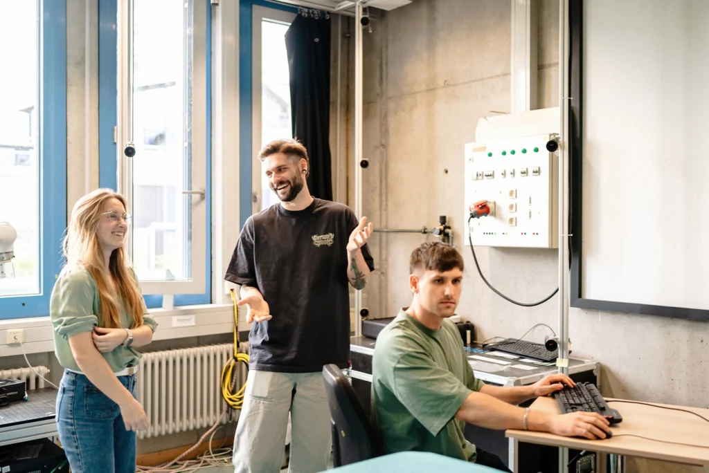 Drei Personen arbeiten und sprechen an Computerarbeitsplätzen in einem technischen Labor mit großen Bildschirmen und Messgeräten.