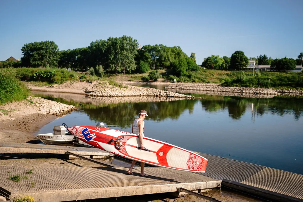 Person mit Sportbekleidung und Sonnenhut trägt ein Surfbrett am Ufer des Neckar in Mannheim bei sonnigem Wetter.