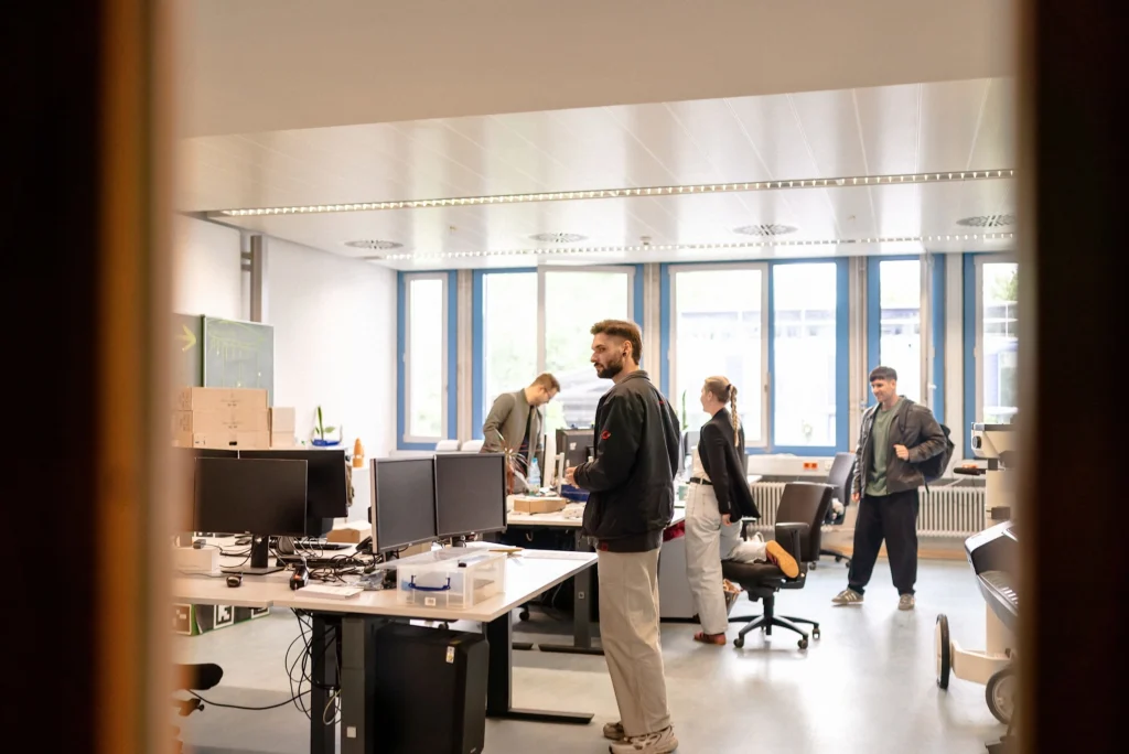 Mehrere Personen stehen und arbeiten in einem Büro mit Schreibtischen, Bildschirmen und großen Fenstern.