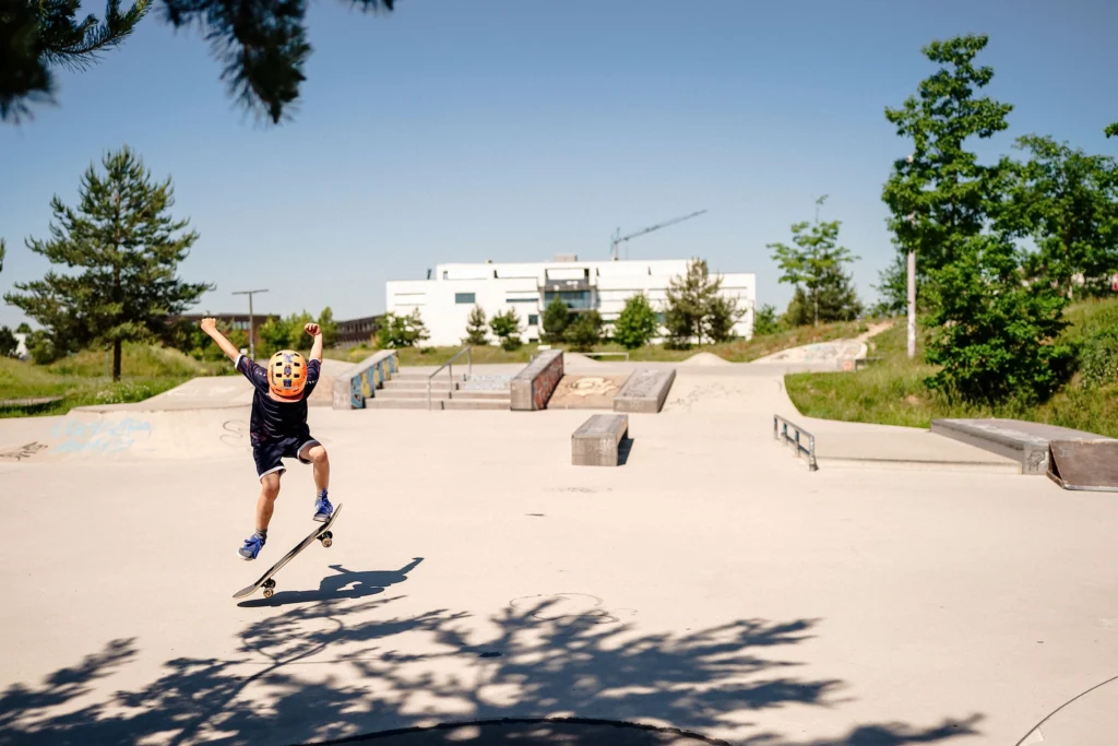 Ein Kind mit Helm springt auf einem Skateboard über eine Rampe in einem Skatepark mit verschiedenen Hindernissen, umgeben von Grünflächen und Bäumen.