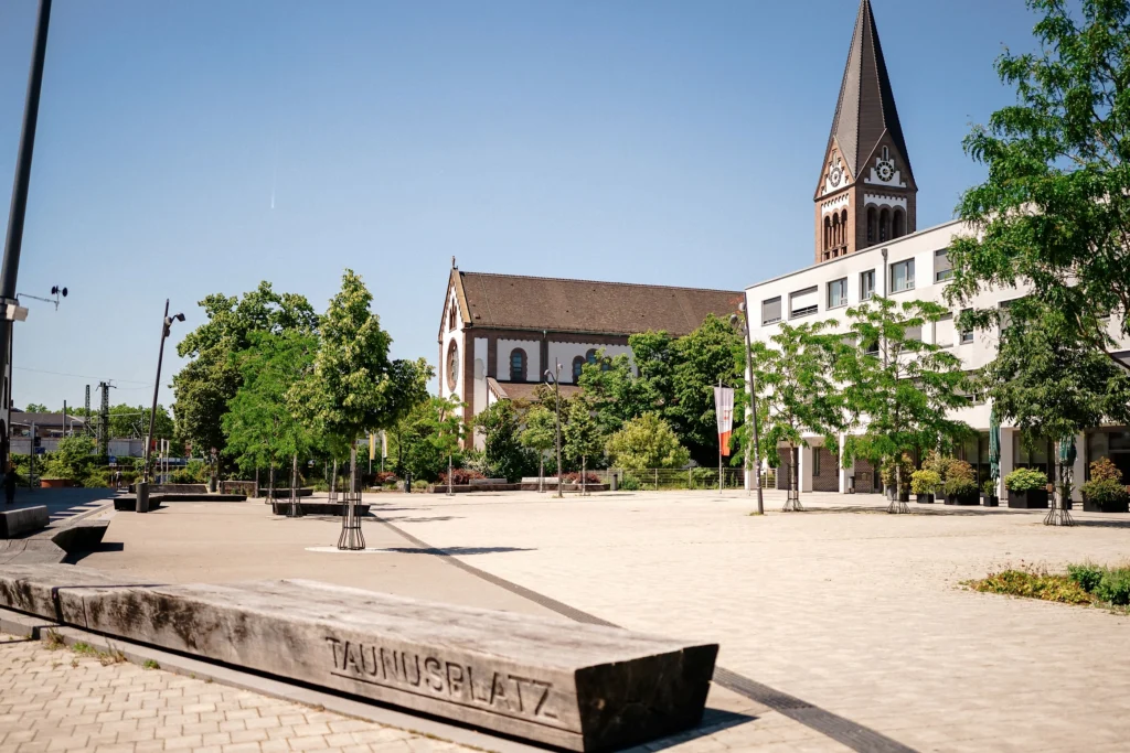 Offener Platz mit Bänken und Bäumen, eine Kirche und weitere Gebäude im Hintergrund.