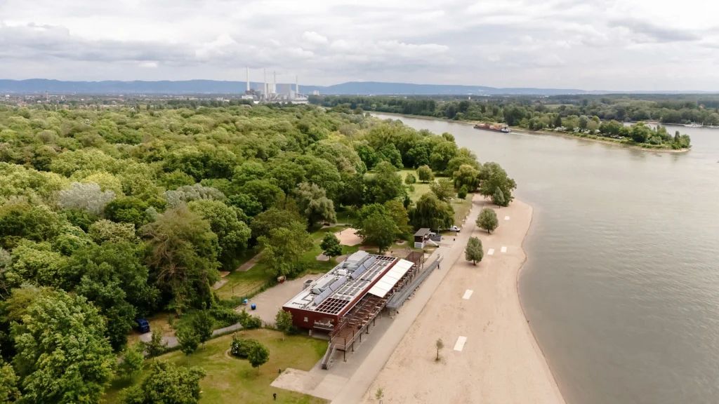 Luftaufnahme des Strandbads Rhein Mannheim mit Sandstrand, Gebäuden und angrenzendem Flussufer, umgeben von Waldflächen.
