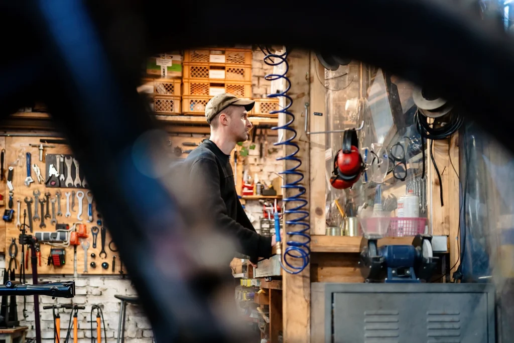 Eine Person steht in einer Fahrradwerkstatt vor einer Werkbank und Werkzeugwand, eingerahmt von unscharfen Fahrradteilen im Vordergrund.