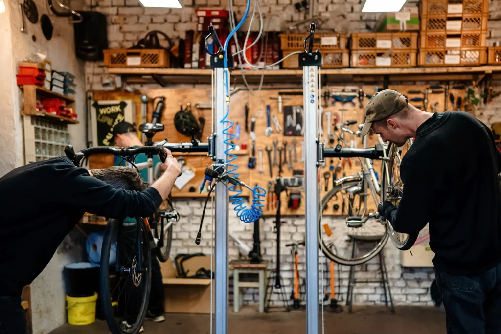 Zwei Personen arbeiten an aufgehängten Fahrrädern in einer gut ausgestatteten Fahrradwerkstatt, deren Wände mit Werkzeugen, Ersatzteilen und Regalen voller Material bestückt sind.