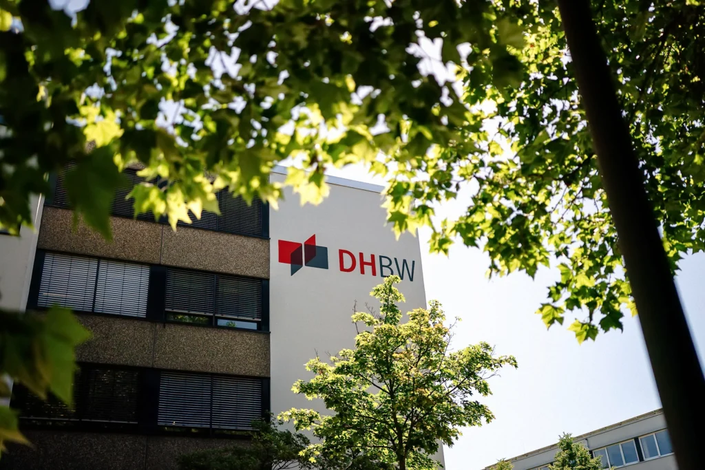 Das Gebäude der Dualen Hochschule Baden-Württemberg ist teilweise von Bäumen verdeckt, während das DHBW-Logo gut sichtbar an der Fassade angebracht ist.