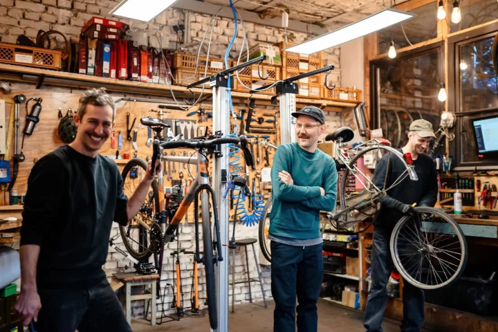 Drei Mitarbeitende stehen lächelnd zwischen Fahrrädern und Werkzeug in einer Fahrradwerkstatt.