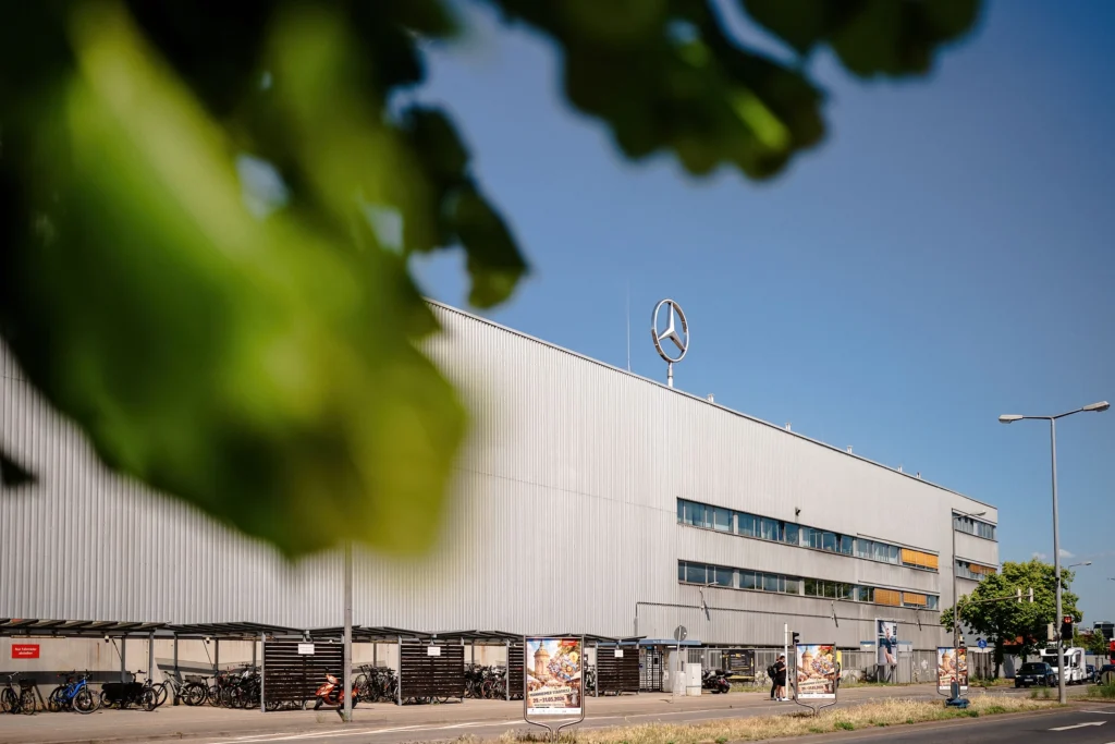 Großes Industriegebäude mit Mercedes-Stern auf dem Dach, im Vordergrund unscharfe Blätter und eine Straße mit Fahrradständern.