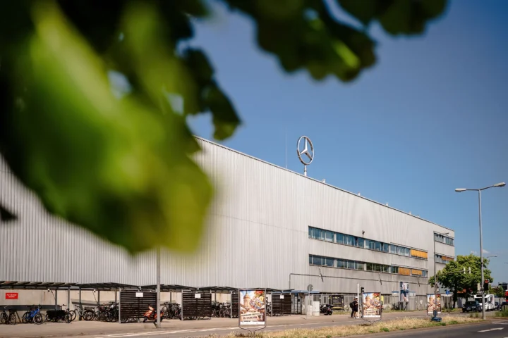 Großes Industriegebäude mit Mercedes-Stern auf dem Dach, im Vordergrund unscharfe Blätter und eine Straße mit Fahrradständern.