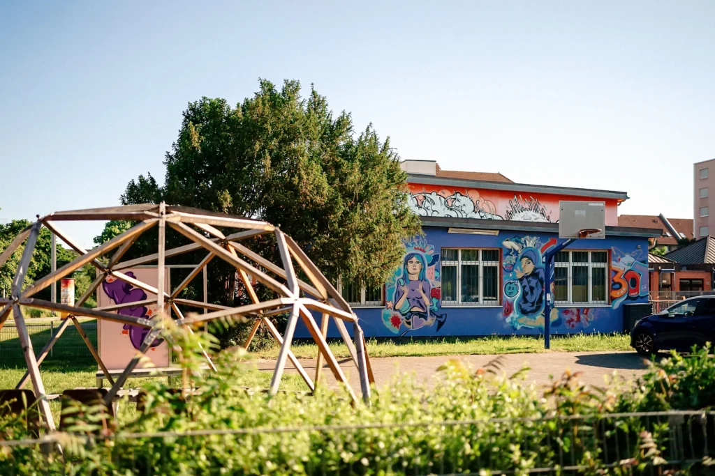 Spielplatz mit Klettergerüst vor einem Gebäude mit bunten Graffiti, umgeben von Grünflächen an einem klaren Tag.