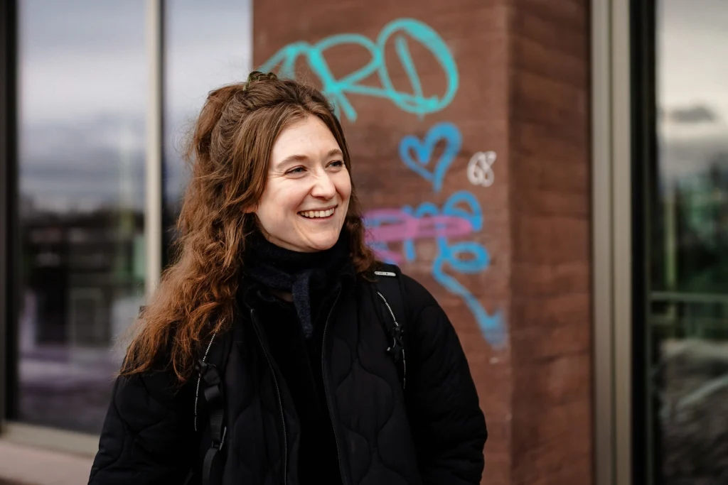 Eine lächelnde Person steht im Freien vor einem Gebäude mit Graffiti im Hintergrund.