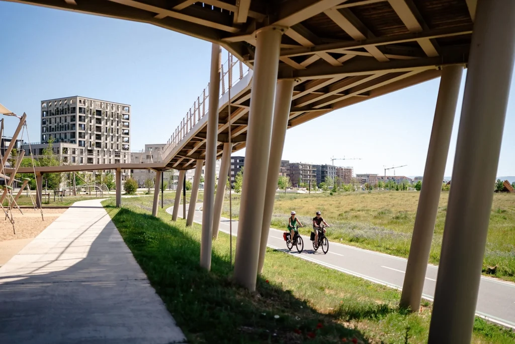 Zwei Radfahrer fahren auf einem Weg hinter einem Klettergerüst in einem Park mit modernen Gebäuden im Hintergrund.