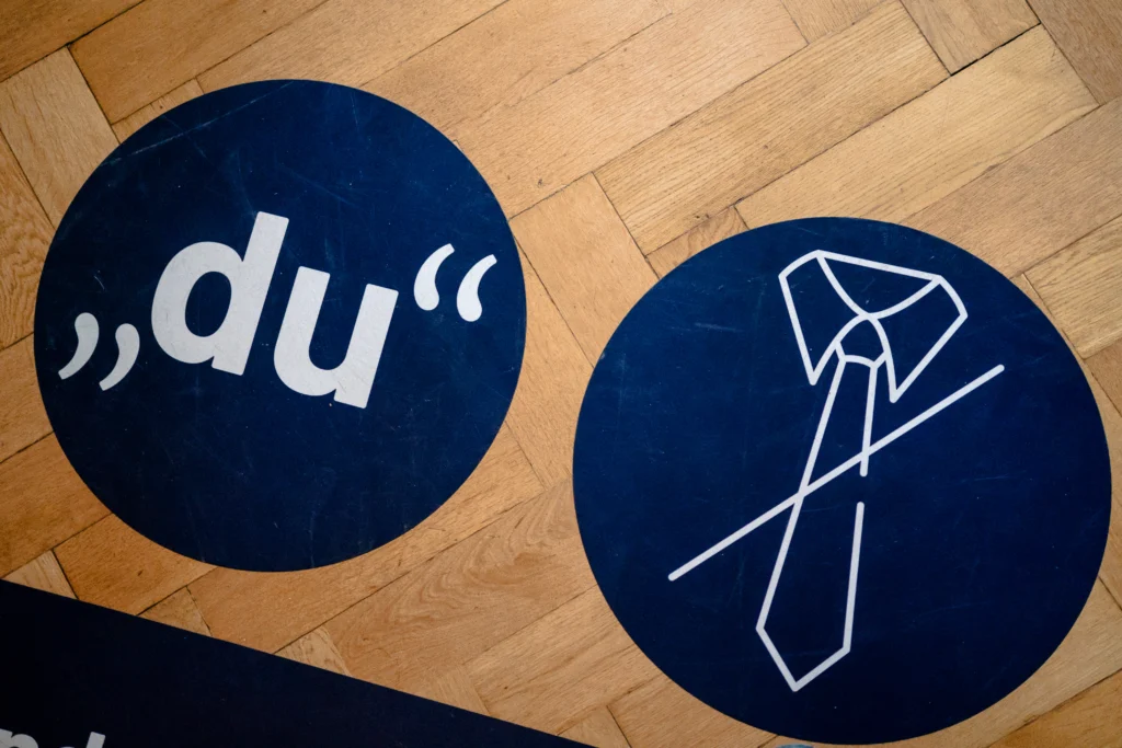 Zwei runde Hinweisschilder auf einem Holzfußboden zeigen die Aufschrift „du“ sowie ein Symbol einer Krawatte.