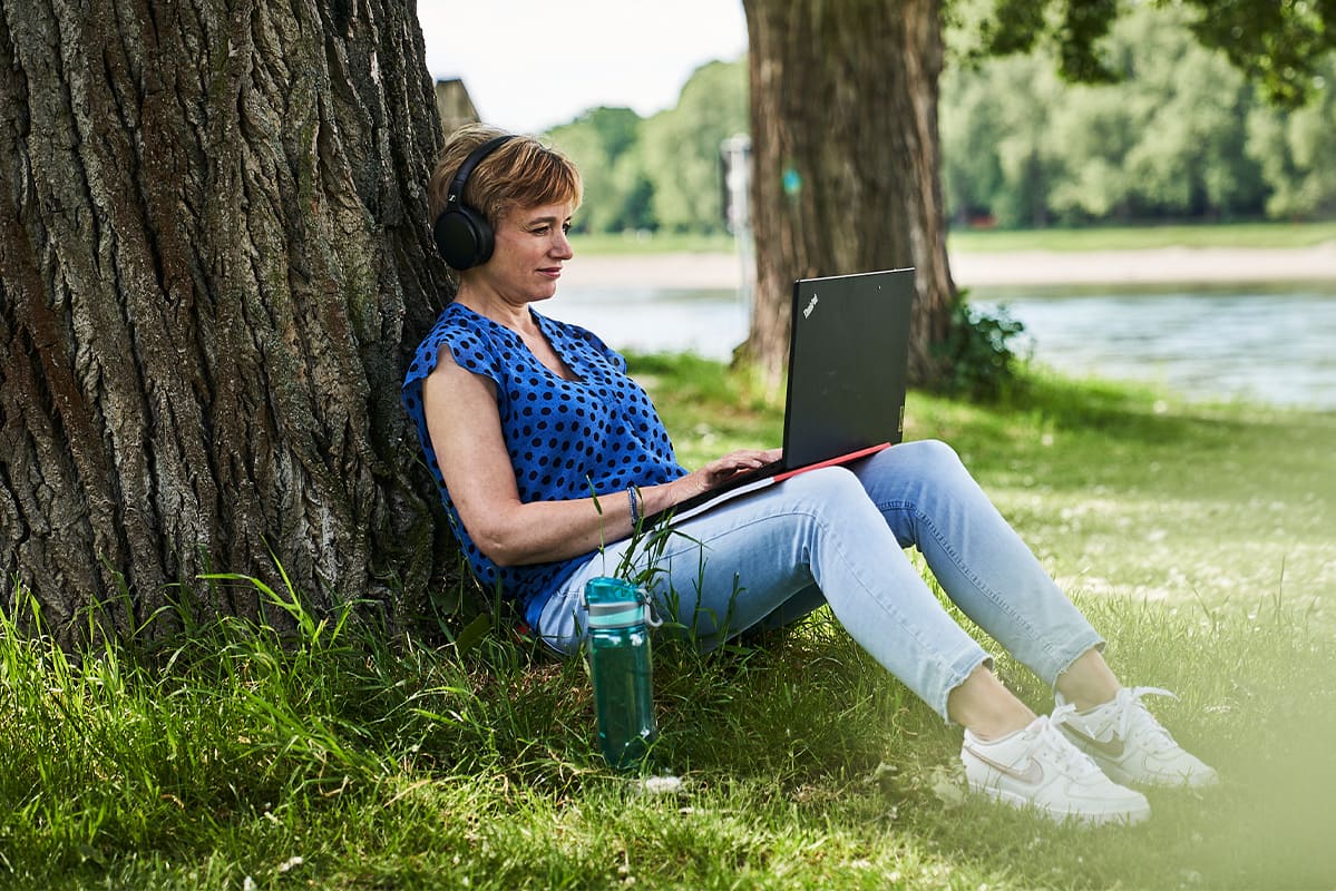 Eine Frau genießt die Ruhe am Rheinufer in Mannheim. Sie sitzt auf dem grünen Rasen und blickt auf ihren Laptop.