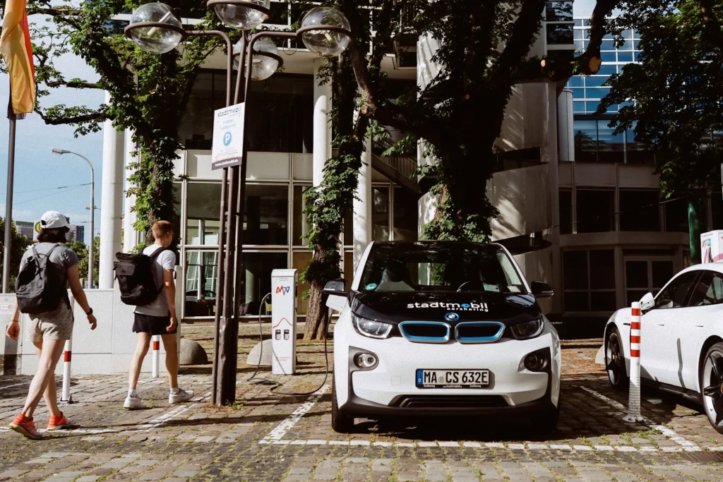 Kleiner weißer Elektro-Kleinwagen mit Aufschrift Stadtmobil steht an einer Ladestation in einer städtischen Umgebung mit zwei Fußgängern im Hintergrund.