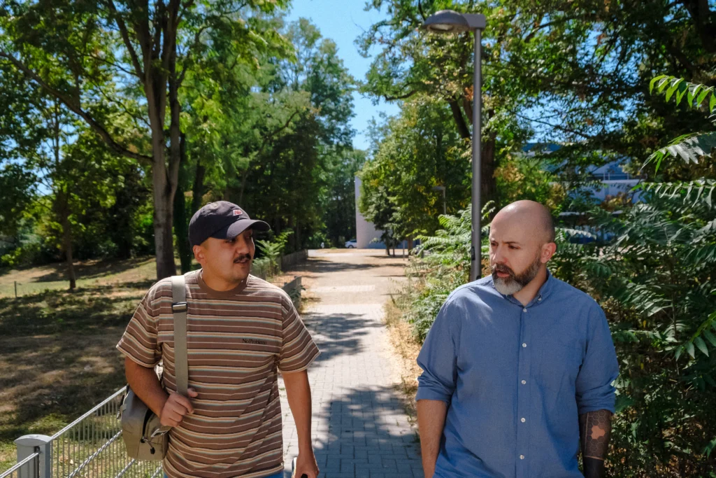 Zwei Personen führen ein Gespräch während eines Spaziergangs durch eine grüne Wohnanlage.