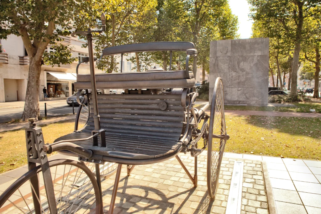 Ein Modell des von Benz entwickelten Automobils steht als Denkmal auf einem Grünstreifen an einer Allee