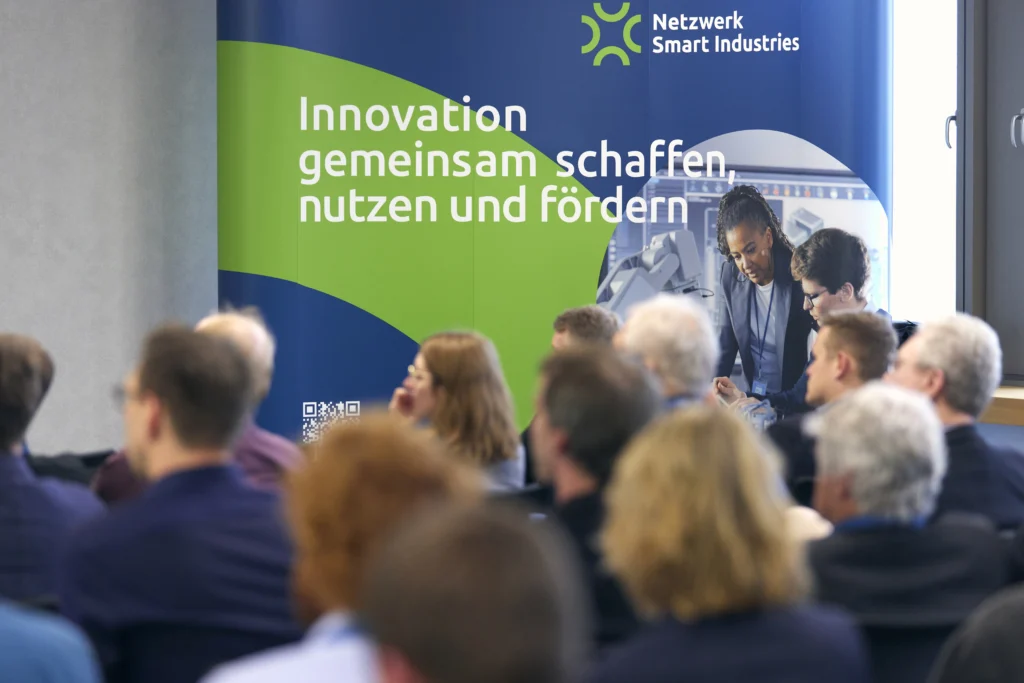 Publikum sitzt vor einem Banner mit dem Text Innovation gemeinsam schaffen, nutzen und fördern und dem Logo Netzwerk Smart Industries.