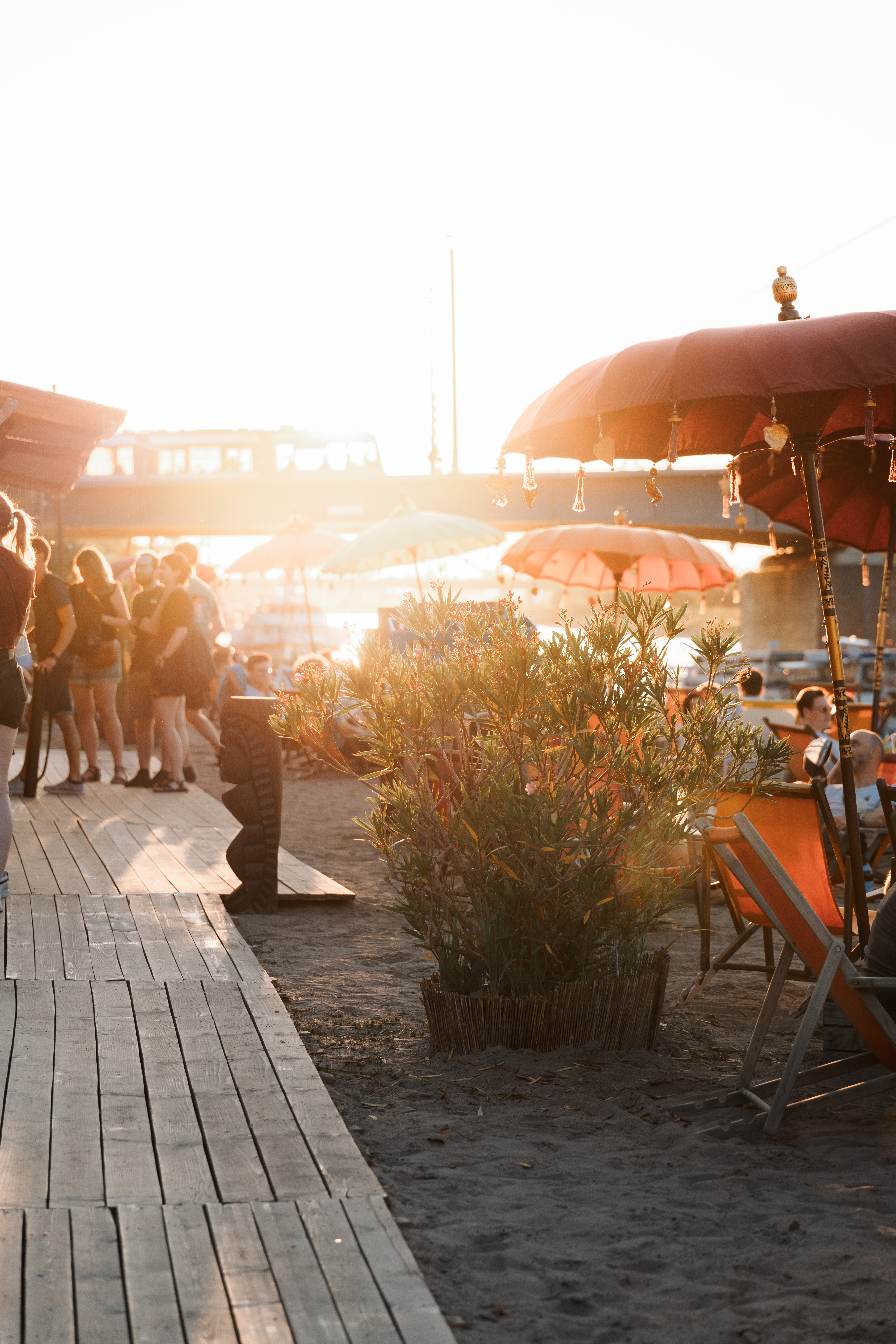Menschen sitzen und stehen auf einem Sandstrand mit Liegestühlen und Sonnenschirmen am Wasser während des Sonnenuntergangs.