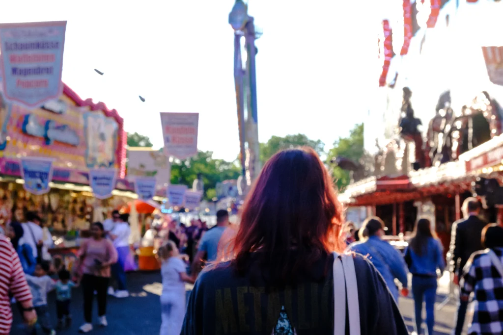 Eine Person geht auf einem belebten Festgelände zwischen Markt- und Fahrgeschäften, während die tief stehende Sonne die Szene beleuchtet.