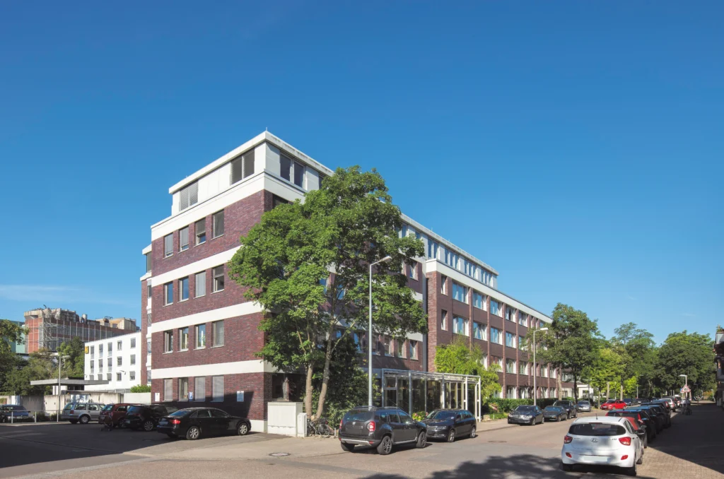 Die Diringer & Scheidel Zentrale, ein Mehrstöckiges Bürogebäude mit rotem Backstein und weißen Fensterrahmen an einer Straße mit parkenden Autos und Bäumen im Vordergrund.