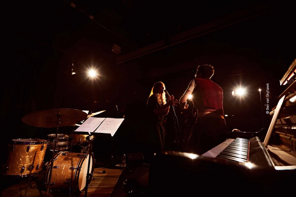 Sängerin und Kontrabassspieler geben Konzert auf beleuchteter Bühne in dunklem Saal. Links von ihnen stehen Schlagzeug und Notenständer, rechts von ihnen ein Klavier.