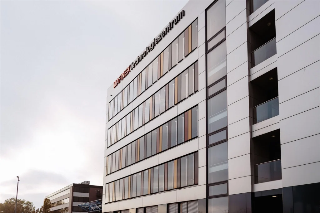 Die moderne Glasfassade des MAFINEX Technologiezentrums in Mannheim erstreckt sich über mehrere Stockwerke und steht vor einem hellen, leicht bewölkten Himmel.