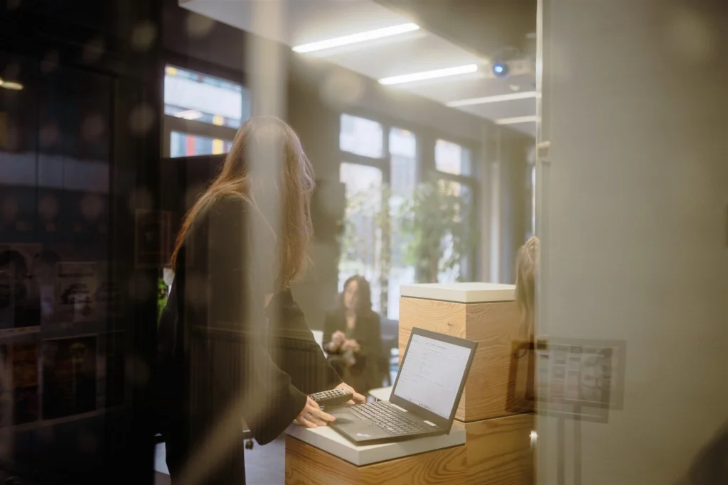 Frau steht an einem Laptop auf einem Holztisch in einem Raum mit großen Fenstern, im Hintergrund sitzt eine weitere Person.