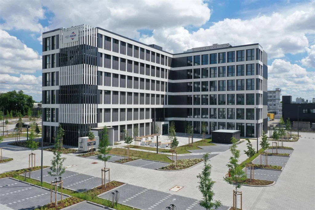 Modernes Bürogebäude mit mehreren Stockwerken und vertikalen Fassadenelementen, umgeben von Parkplätzen und jungen Bäumen auf dem Gelände der Fuchs SE Zentrale.