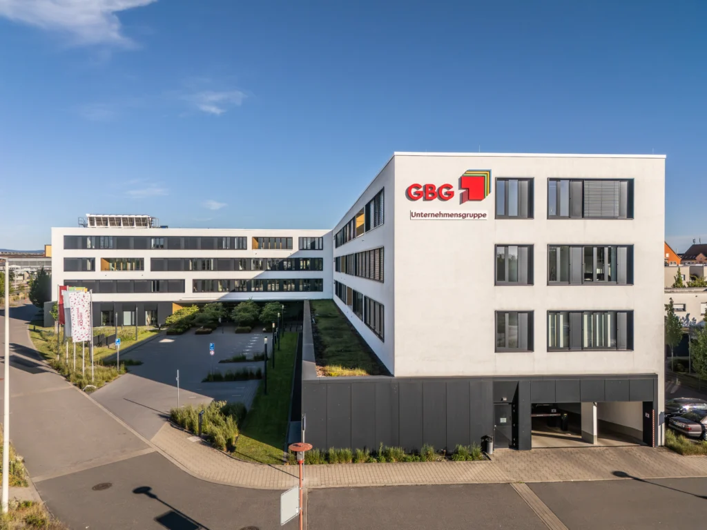 Modernes Bürogebäude mit dem Schriftzug 'GBG Unternehmensgruppe' an der Fassade und mehreren Fenstern, umgeben von einem Parkplatz und Grünflächen.